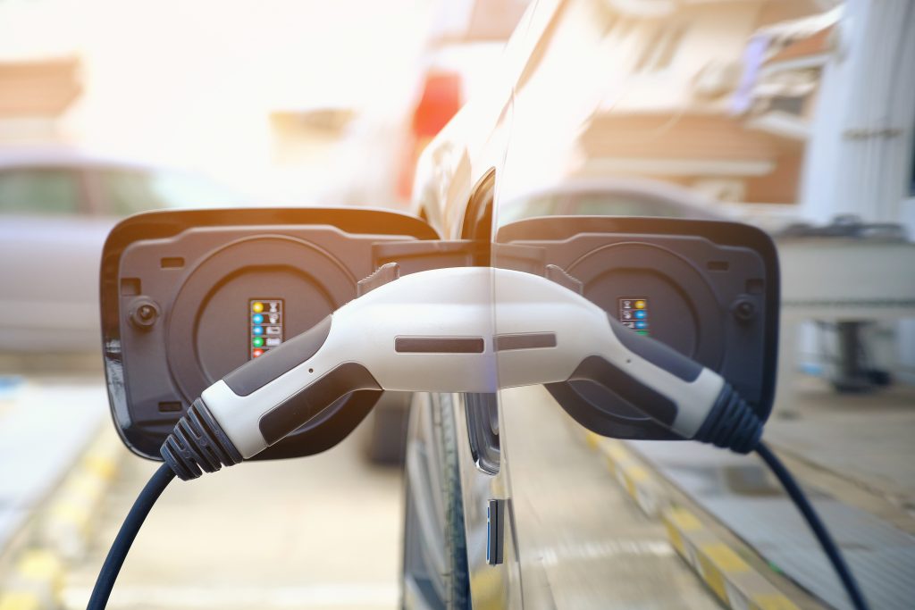 Electric vehicle plugged into a charging station, with one connector inserted into the charging port and its reflection visible on the car body.